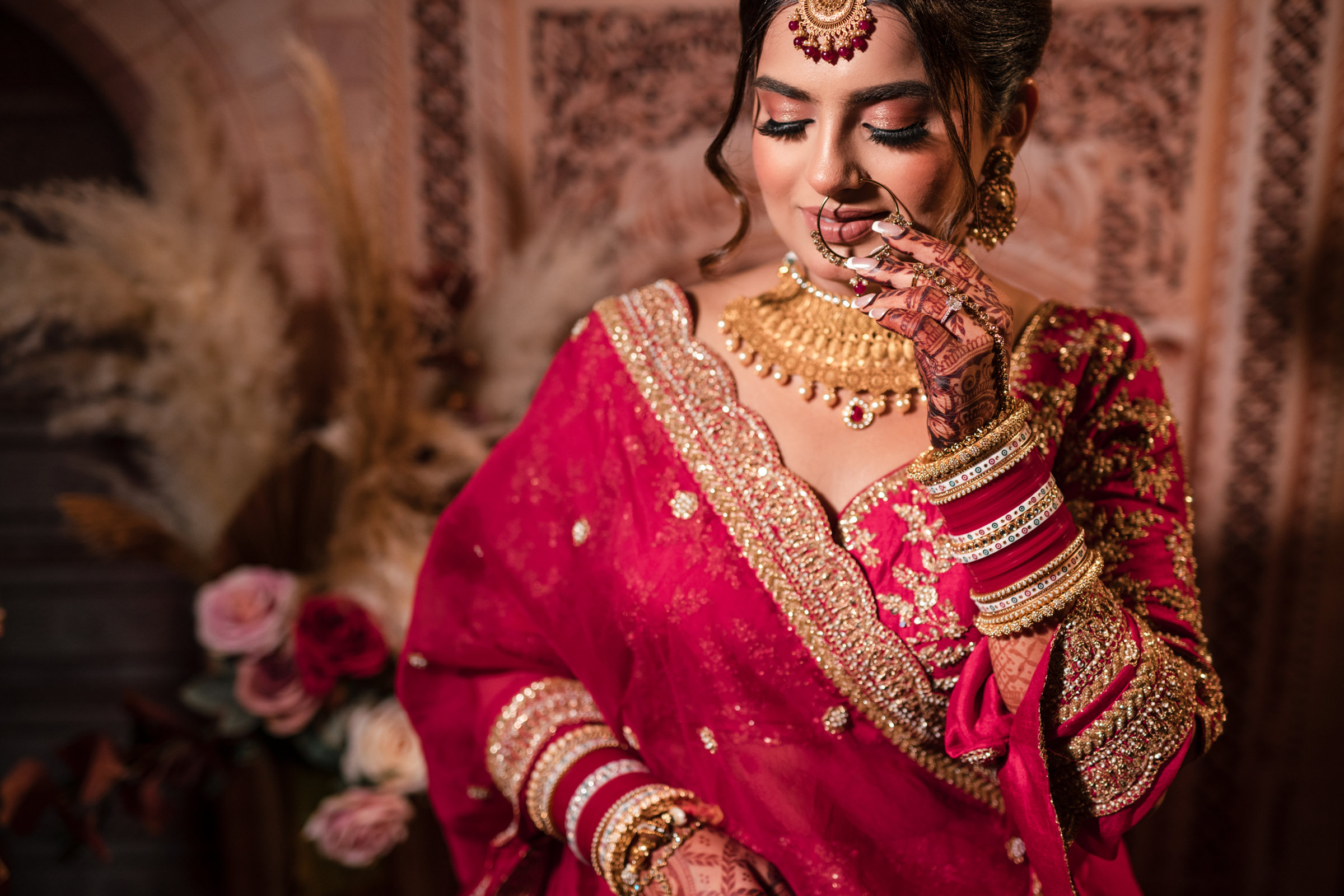 Bride in a red and gold lehenga with mehndi and chooda, smiling with eyes lowered